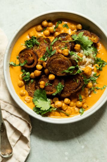 Overhead shot of a bowl of eggplant curry with roasted chickpeas over rice, topped with crispy eggplant rounds and fresh cilantro leaves, curry sauce a vibrant orange-red.