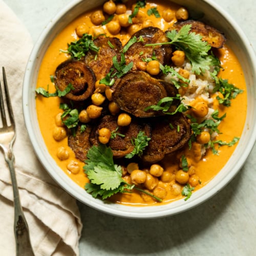 Overhead shot of a bowl of eggplant curry with roasted chickpeas over rice, topped with crispy eggplant rounds and fresh cilantro leaves, curry sauce a vibrant orange-red.