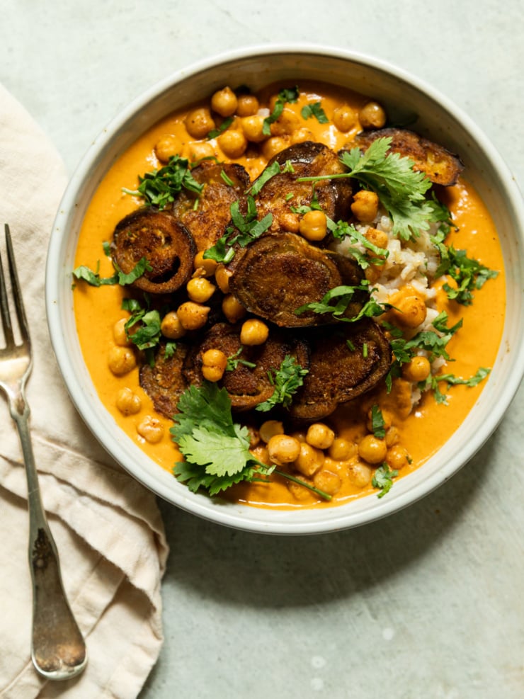 Overhead shot of a bowl of eggplant curry with roasted chickpeas over rice, topped with crispy eggplant rounds and fresh cilantro leaves, curry sauce a vibrant orange-red.