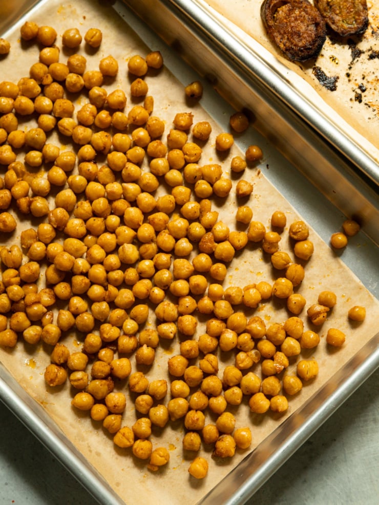 Roasted chickpeas spread on a parchment-lined baking sheet, golden and lightly golden at the edges, with roasted eggplant halves visible on a second tray in the corner.