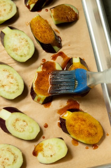 Raw eggplant slices on a parchment-lined baking sheet being brushed with a spiced oil mixture using a blue silicone pastry brush.