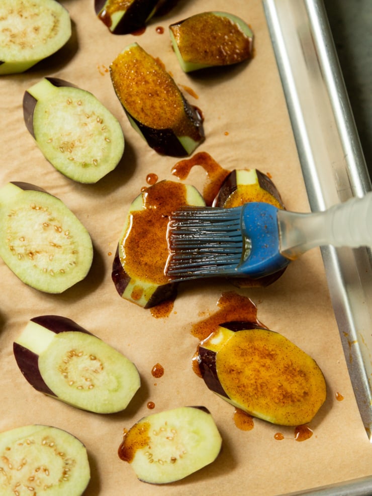 Raw eggplant slices on a parchment-lined baking sheet being brushed with a spiced oil mixture using a blue silicone pastry brush.