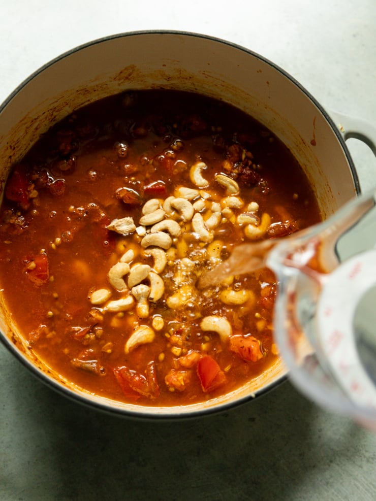 Water being poured into a pot of simmering tomato sauce with raw cashews, mid-preparation for the curry base.