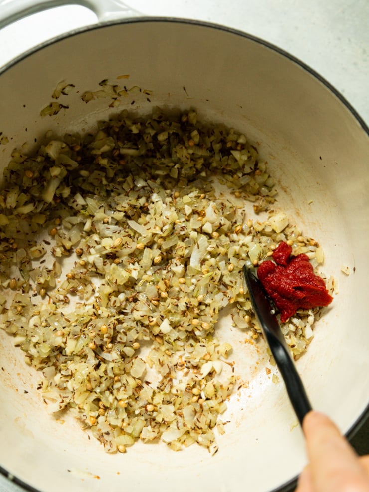 Sautéed onions, garlic, and whole spices in a Dutch oven, with a dollop of tomato paste being stirred in with a spatula.