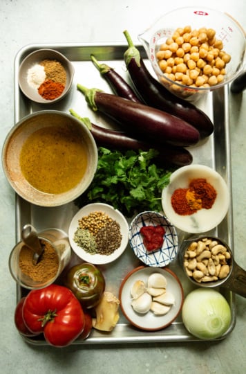 Overhead flat-lay of raw ingredients for the curry: whole eggplants, chickpeas, cashews, tomatoes, onion, garlic, ginger, cilantro, and small bowls of ground and whole spices arranged on a metal tray.