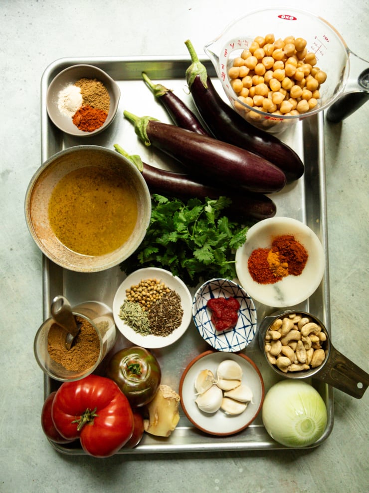 Overhead flat-lay of raw ingredients for the curry: whole eggplants, chickpeas, cashews, tomatoes, onion, garlic, ginger, cilantro, and small bowls of ground and whole spices arranged on a metal tray.