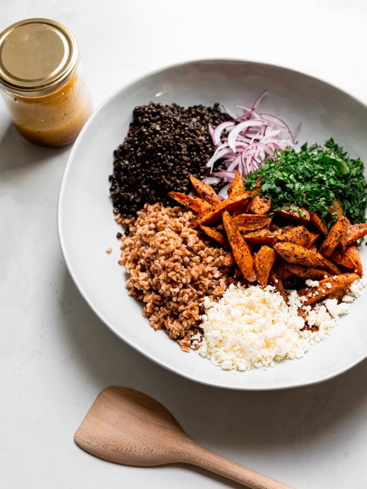 Cooked black lentils, farro, roasted carrots, red onion, chopped parsley and dill, and crumbled vegan feta arranged in sections in a white bowl, with a jar of dressing beside it.