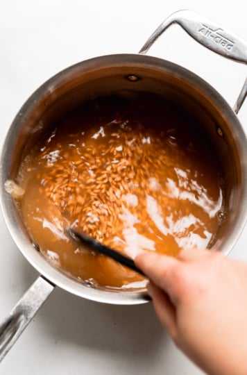 A hand stirring farro in simmering apple cider mixture in a stainless steel saucepan.