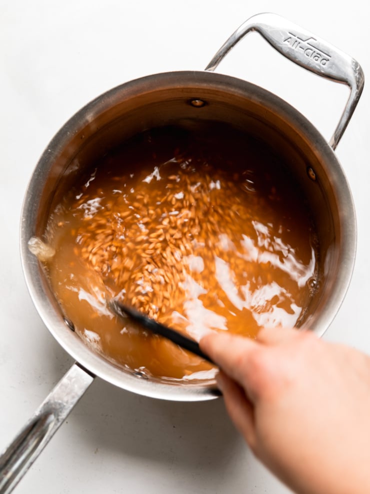 A hand stirring farro in simmering apple cider mixture in a stainless steel saucepan.
