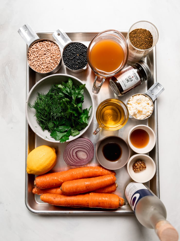 Ingredients for farro lentil salad on a tray: farro, black lentils, broth, za'atar, black lime, parsley, dill, olive oil, crumbled vegan feta, lemon, red onion, carrots and dressing ingredients.