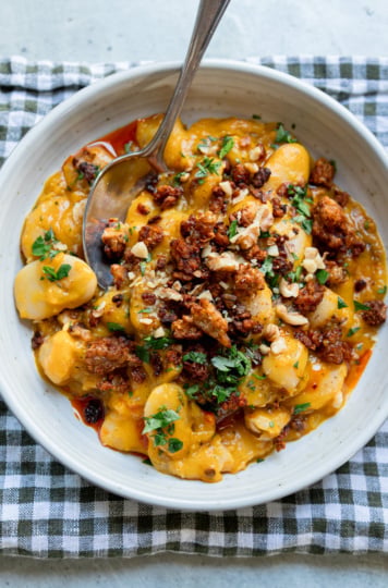 Creamy butter beans in a golden honey nut squash sauce, topped with crispy smoky tempeh crumbles, chopped walnuts, fresh parsley and a drizzle of chili oil, served in a speckled white bowl with a spoon on a green gingham napkin.