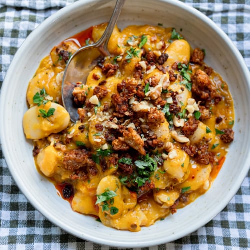 Creamy butter beans in a golden honey nut squash sauce, topped with crispy smoky tempeh crumbles, chopped walnuts, fresh parsley and a drizzle of chili oil, served in a speckled white bowl with a spoon on a green gingham napkin.