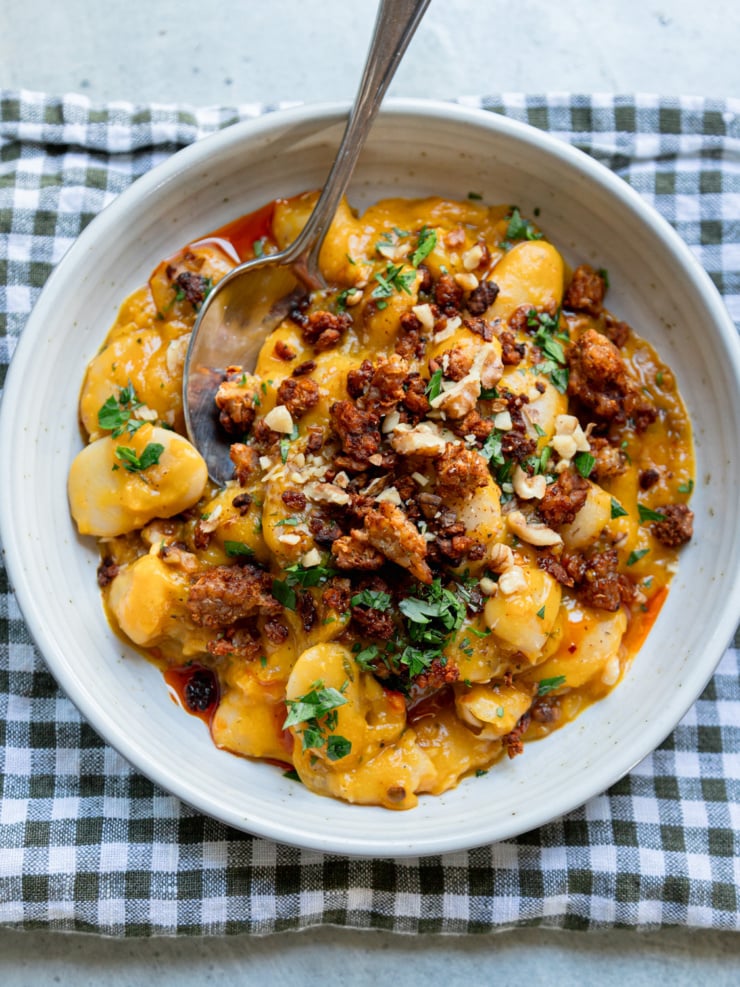 Creamy butter beans in a golden honey nut squash sauce, topped with crispy smoky tempeh crumbles, chopped walnuts, fresh parsley and a drizzle of chili oil, served in a speckled white bowl with a spoon on a green gingham napkin.