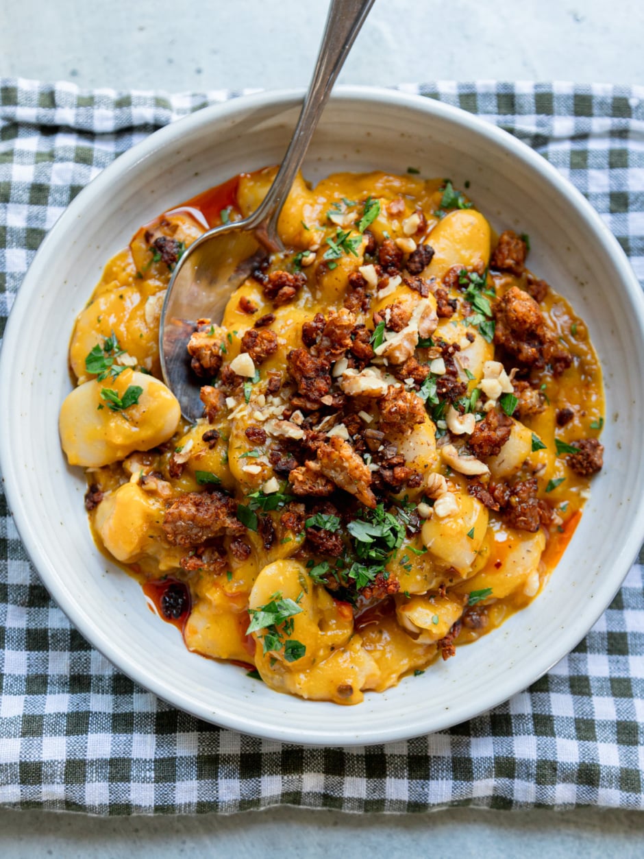 Creamy butter beans in a golden honey nut squash sauce, topped with crispy smoky tempeh crumbles, chopped walnuts, fresh parsley and a drizzle of chili oil, served in a speckled white bowl with a spoon on a green gingham napkin.