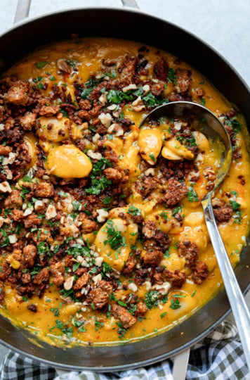 A large skillet of butter beans in roasted garlic and honey nut squash sauce, covered with smoky roasted tempeh bits, chopped walnuts and parsley, with a serving ladle resting in the pan.