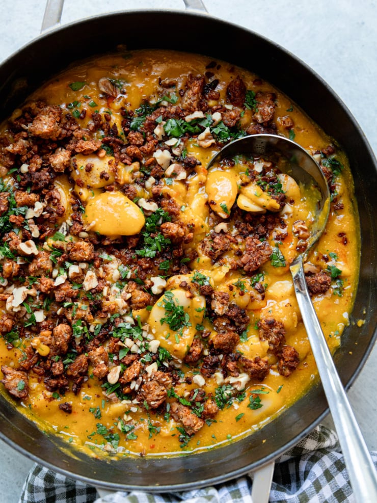 A large skillet of butter beans in roasted garlic and honey nut squash sauce, covered with smoky roasted tempeh bits, chopped walnuts and parsley, with a serving ladle resting in the pan.