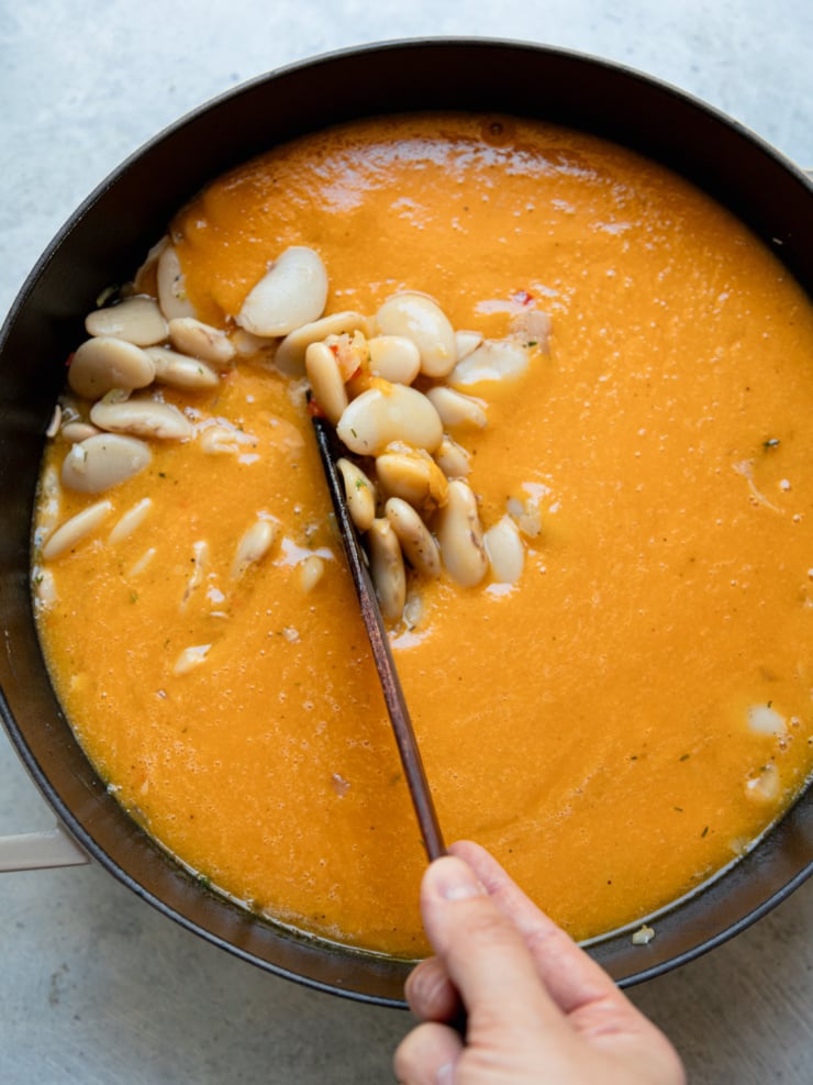 A hand stirring cooked butter beans into a smooth, bright orange honey nut squash sauce in a dark skillet with a wooden spoon.