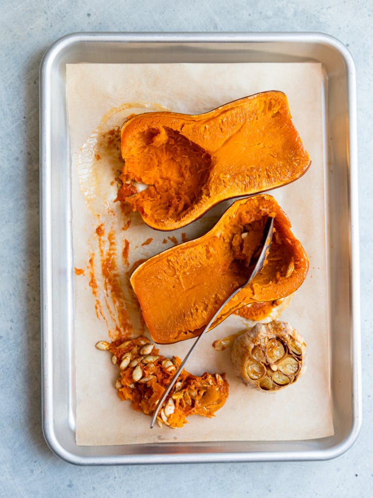 Two roasted honey nut squash halves on a parchment-lined sheet pan, with a spoon scooping out the seeds beside a head of soft, caramelized roasted garlic.