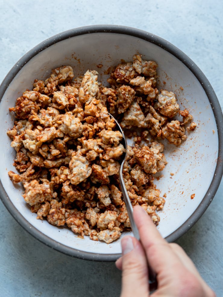 A hand stirring crumbled tempeh with smoky spices in a white bowl before roasting.