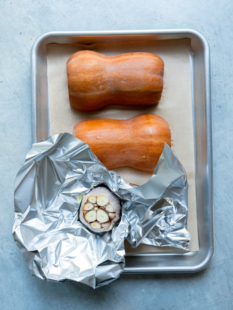 Honey nut squash halves placed cut side down on a parchment-lined sheet pan, next to a head of garlic with its top sliced off, wrapped in foil.