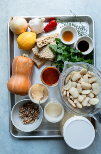 Ingredients for smoky butter beans with honey nut squash sauce on a sheet pan: a honey nut squash, onion, garlic, red chili, lemon, tempeh, fresh parsley and thyme, walnuts, cooked butter beans in a measuring cup, a jar of broth and small dishes of spices and seasonings.
