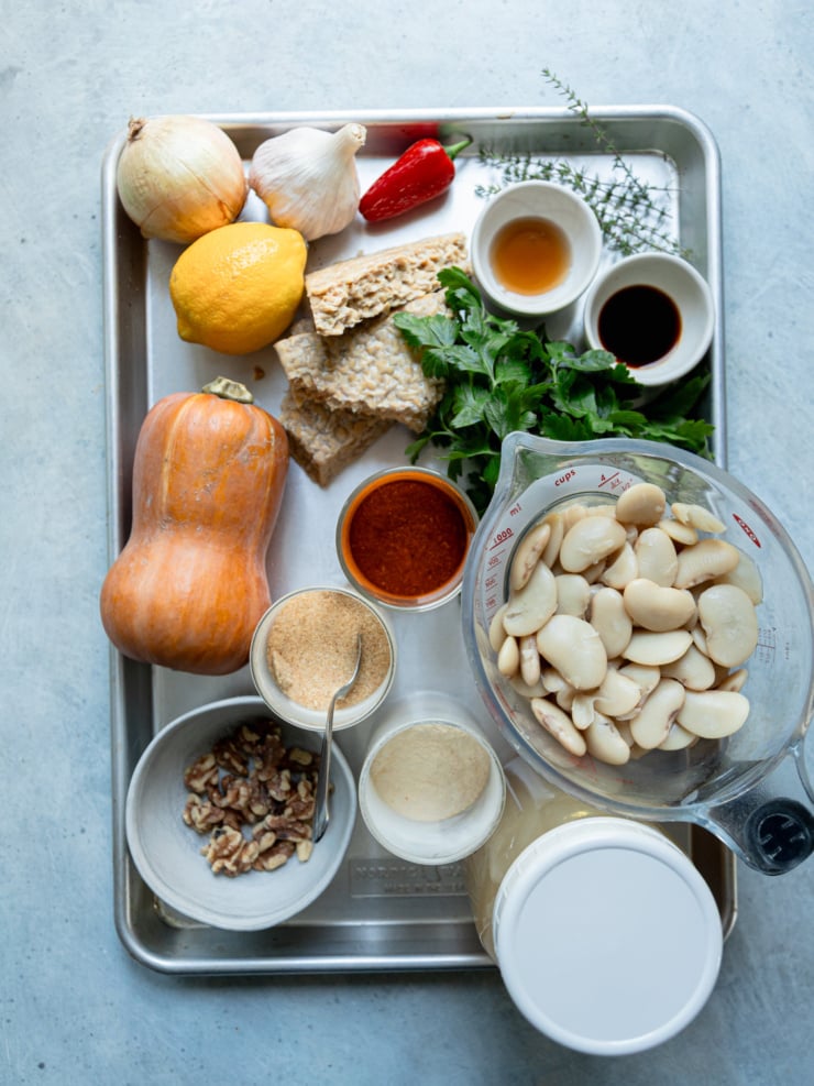 Ingredients for smoky butter beans with honey nut squash sauce on a sheet pan: a honey nut squash, onion, garlic, red chili, lemon, tempeh, fresh parsley and thyme, walnuts, cooked butter beans in a measuring cup, a jar of broth and small dishes of spices and seasonings.