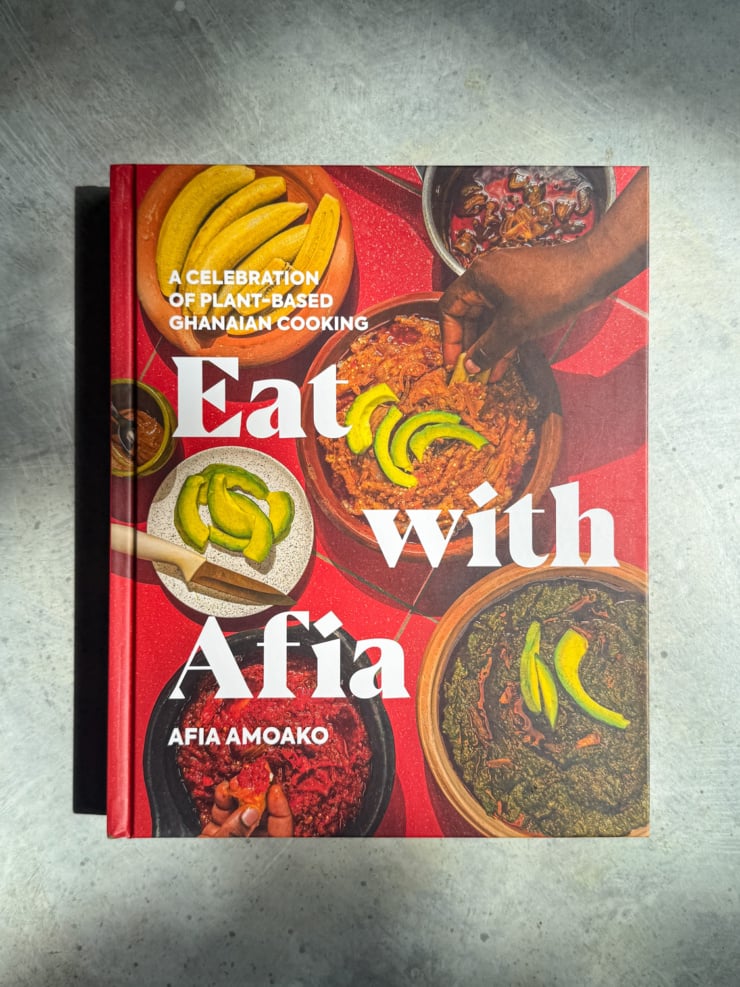 Cover of the cookbook "Eat with Afia: A Celebration of Plant-Based Ghanaian Cooking" by Afia Amoako, showing an overhead spread of dishes including a stew mixture topped with avocado, plantain, stewed greens, and more.