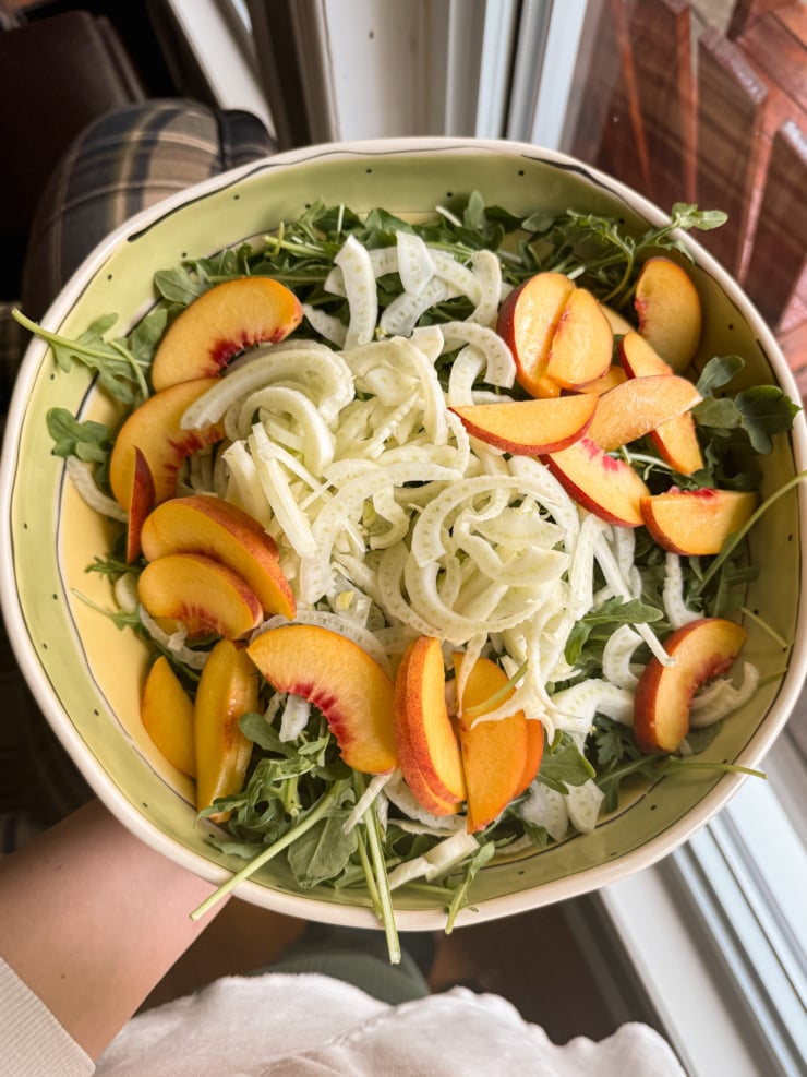Overhead shot of a hand holding a bowl of arugula salad topped with shaved fennel and sliced fresh peaches, near a sunlit window.