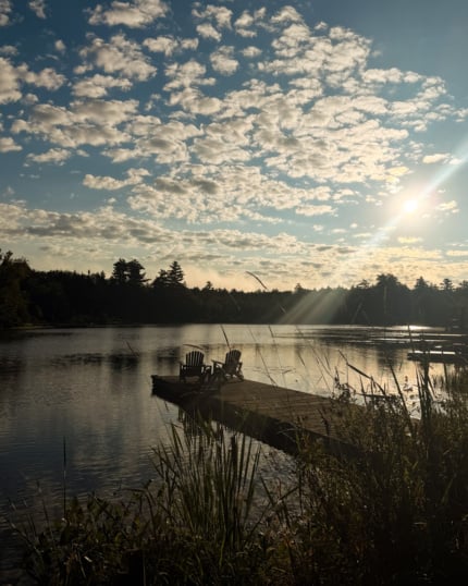 Two Muskoka chairs sitting on a wooden dock extending into a calm lake, under a sky filled with scattered clouds and early morning sun rays, framed by tall grasses in the foreground and tree-lined shores in the distance.