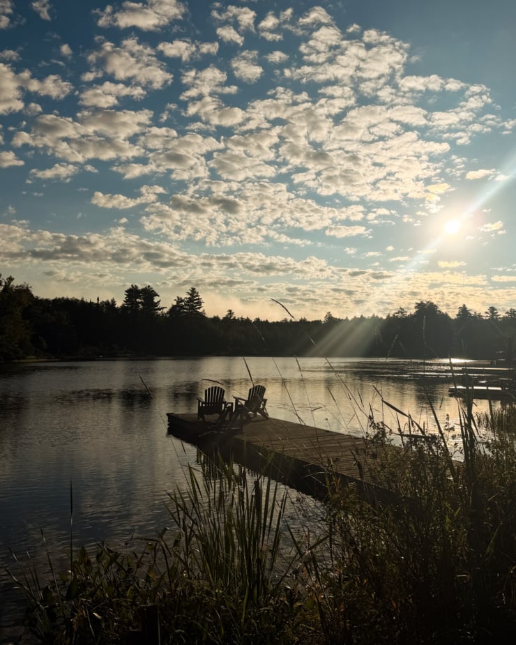 Two Muskoka chairs sitting on a wooden dock extending into a calm lake, under a sky filled with scattered clouds and early morning sun rays, framed by tall grasses in the foreground and tree-lined shores in the distance.
