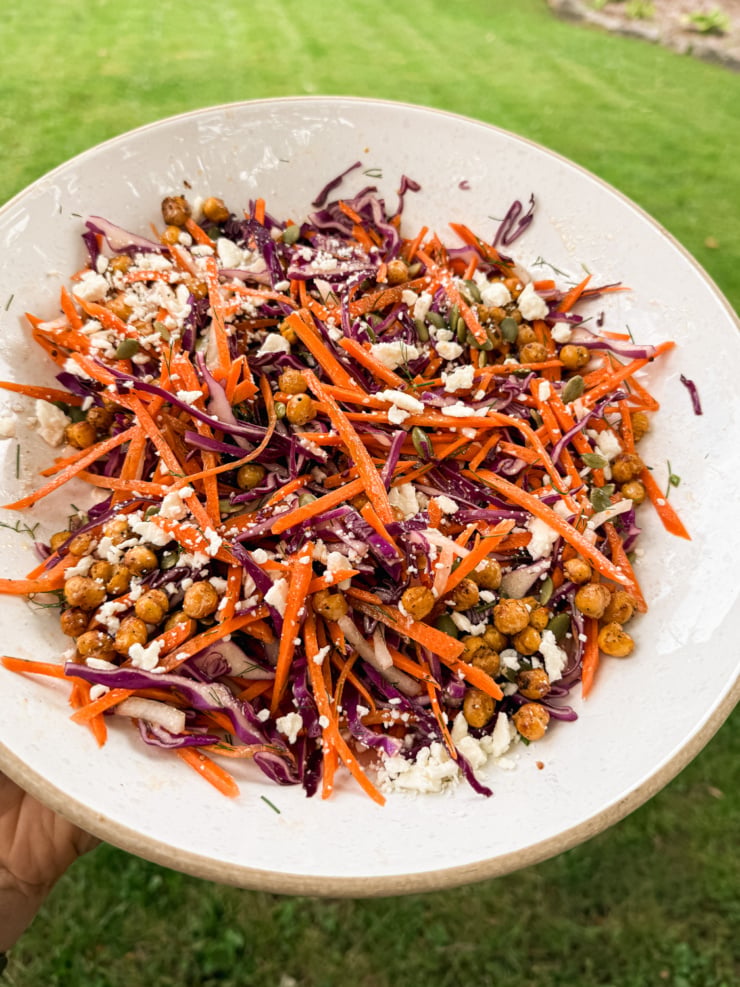 Overhead shot of a chopped salad in a white ceramic bowl, held over a green lawn, with shredded purple cabbage, julienned carrots, crispy spiced chickpeas, crumbled vegan feta, pumpkin seeds, and fresh dill.