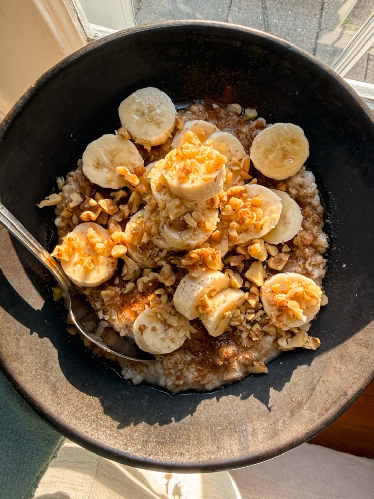A black bowl of oatmeal and barley topped with sliced banana, chopped walnuts, a sprinkle of cinnamon and a drizzle of syrup, lit by warm morning sun beside a window.