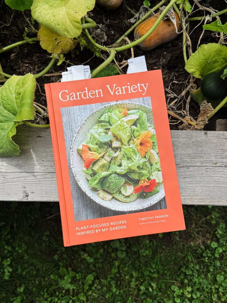 An overhead shot shows the cookbook "Garden Variety" by Timothy Pakron on the ledge of a raised veggie garden bed. Honey nut squashes on vines are seen nearby.