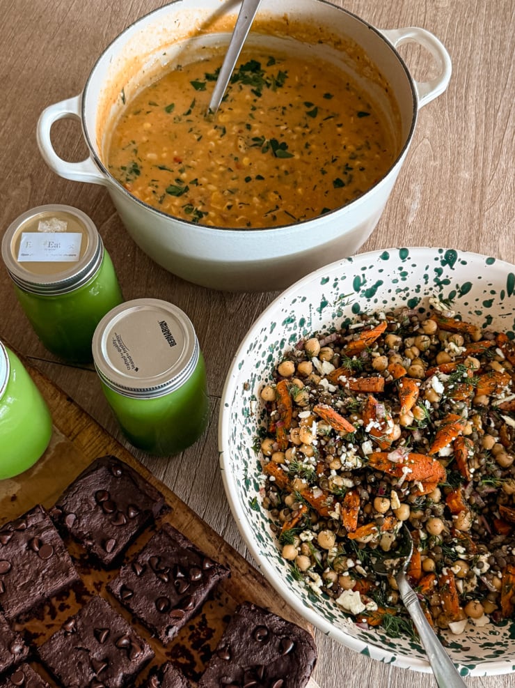 Overhead shot of a meal prep spread with a pot of corn chowder, a bowl of lentil and chickpea salad with roasted carrots and vegan feta, black bean brownies, and three jars of green juice.