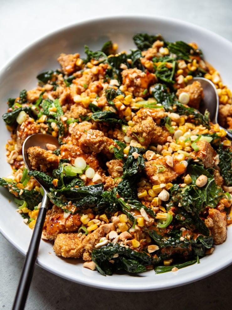Overhead close-up of peanut kale salad with charred corn, crispy tofu bits, and sliced scallions in a white bowl with two serving spoons.