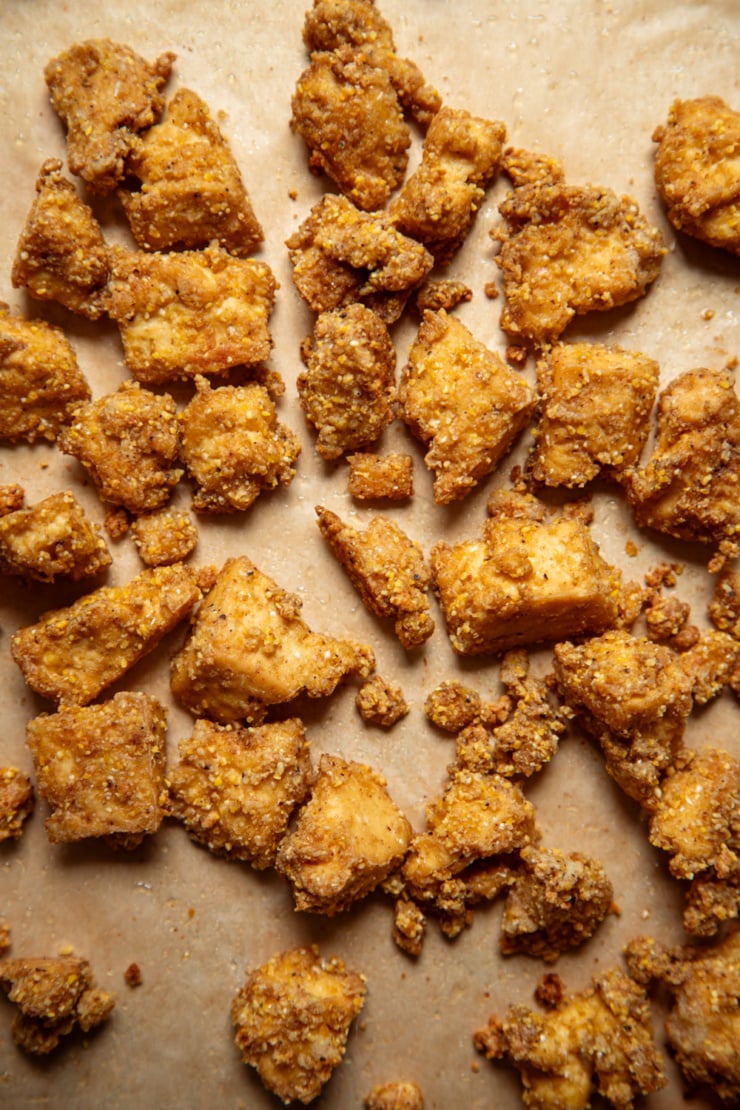 Crispy cornmeal, spice and starch-crusted tofu bits scattered on a parchment-lined baking sheet after roasting.