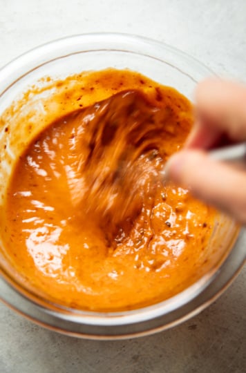Close-up of spicy chili crisp peanut sauce being whisked in a glass bowl, showing its smooth, glossy orange-red texture.