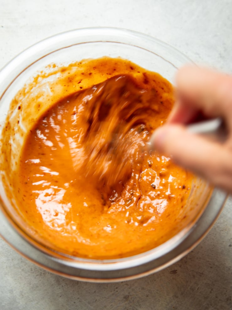 Close-up of spicy chili crisp peanut sauce being whisked in a glass bowl, showing its smooth, glossy orange-red texture.
