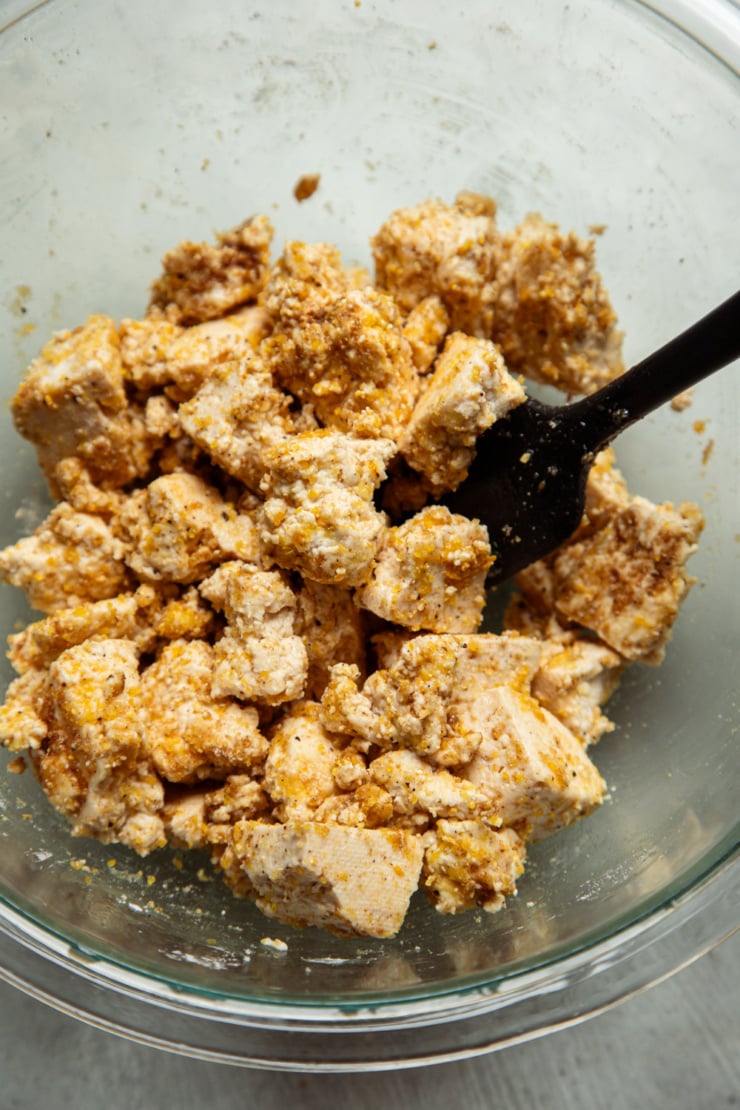 Torn tofu pieces being tossed with cornmeal, arrowroot and seasoning in a glass bowl, coating each piece before baking.
