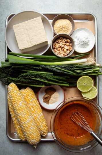 Overhead flat lay of peanut kale salad ingredients, including tofu, corn on the cob, kale, green onions, peanuts, spices, arrowroot, cornmeal, lime, and peanut sauce, arranged on a metal tray.