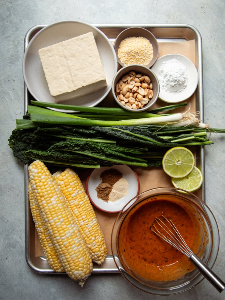 Overhead flat lay of peanut kale salad ingredients, including tofu, corn on the cob, kale, green onions, peanuts, spices, arrowroot, cornmeal, lime, and peanut sauce, arranged on a metal tray.