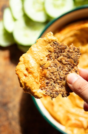 A hand holds a seeded cracker loaded with buffalo butternut squash dip over the bowl, with sliced cucumbers in the background.