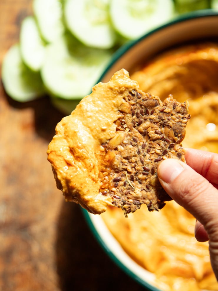 A hand holds a seeded cracker loaded with buffalo butternut squash dip over the bowl, with sliced cucumbers in the background.