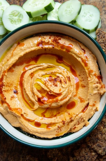 Overhead shot of butternut squash dip swirled in a green-rimmed bowl, topped with olive oil, hot sauce, and paprika, with cucumber slices and seeded crackers alongside.