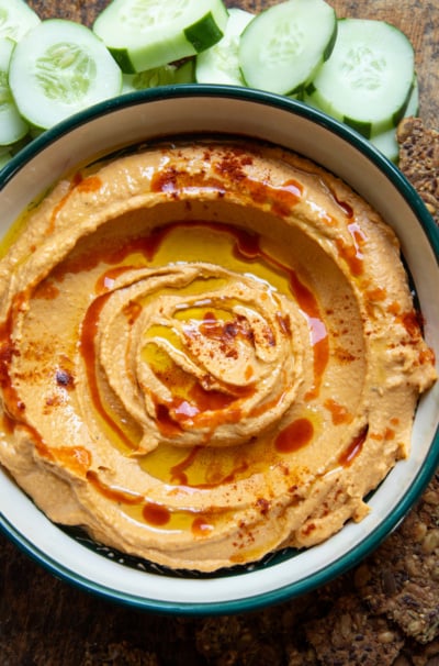 Overhead shot of butternut squash dip swirled in a green-rimmed bowl, topped with olive oil, hot sauce, and paprika, with cucumber slices and seeded crackers alongside.