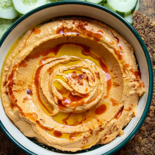 Overhead shot of butternut squash dip swirled in a green-rimmed bowl, topped with olive oil, hot sauce, and paprika, with cucumber slices and seeded crackers alongside.