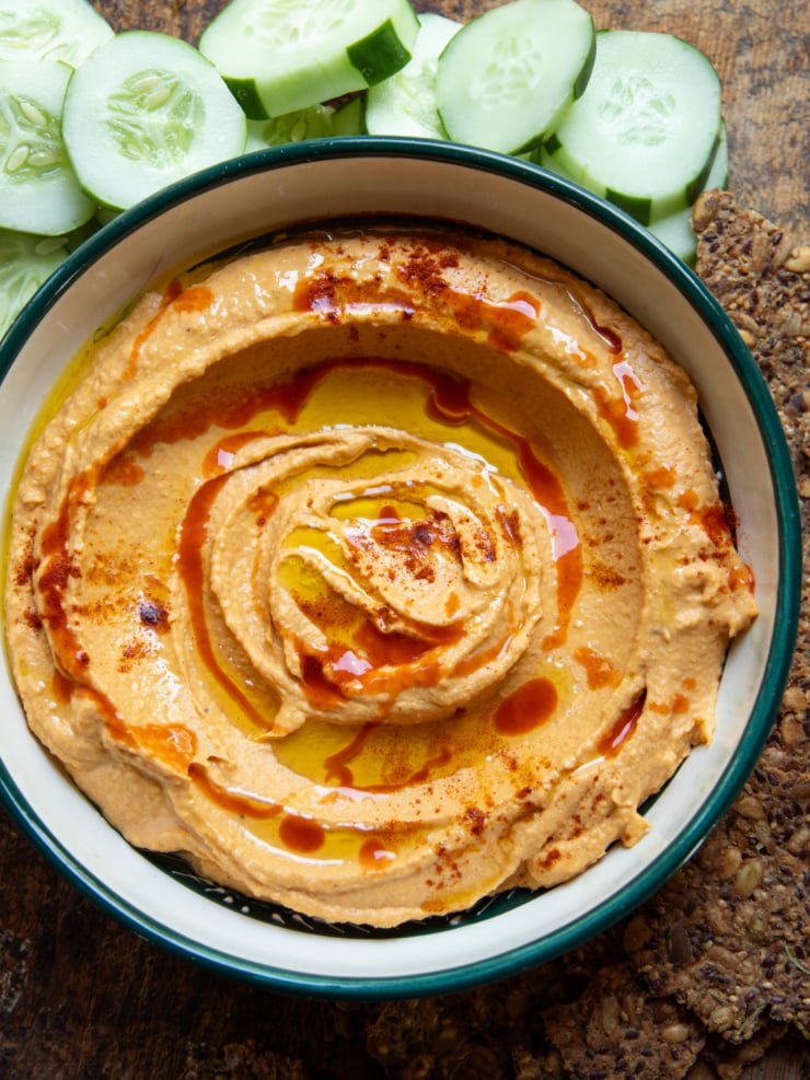 Overhead shot of butternut squash dip swirled in a green-rimmed bowl, topped with olive oil, hot sauce, and paprika, with cucumber slices and seeded crackers alongside.