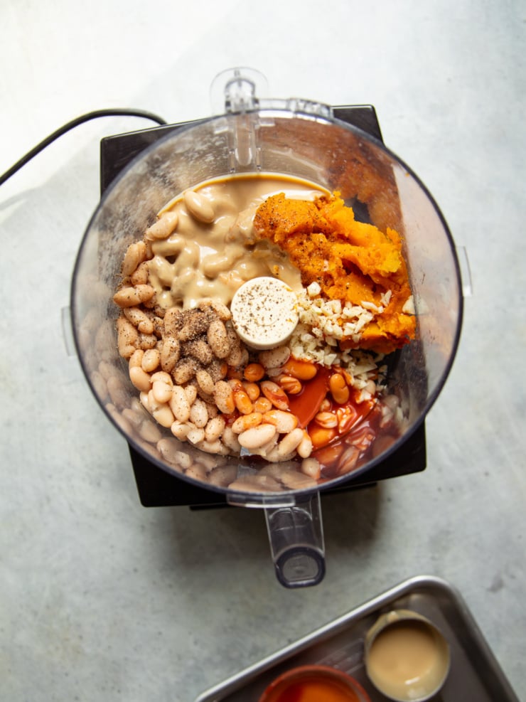 Overhead shot of white beans, roasted squash, tahini, minced garlic, hot sauce, and black pepper in a food processor before blending.