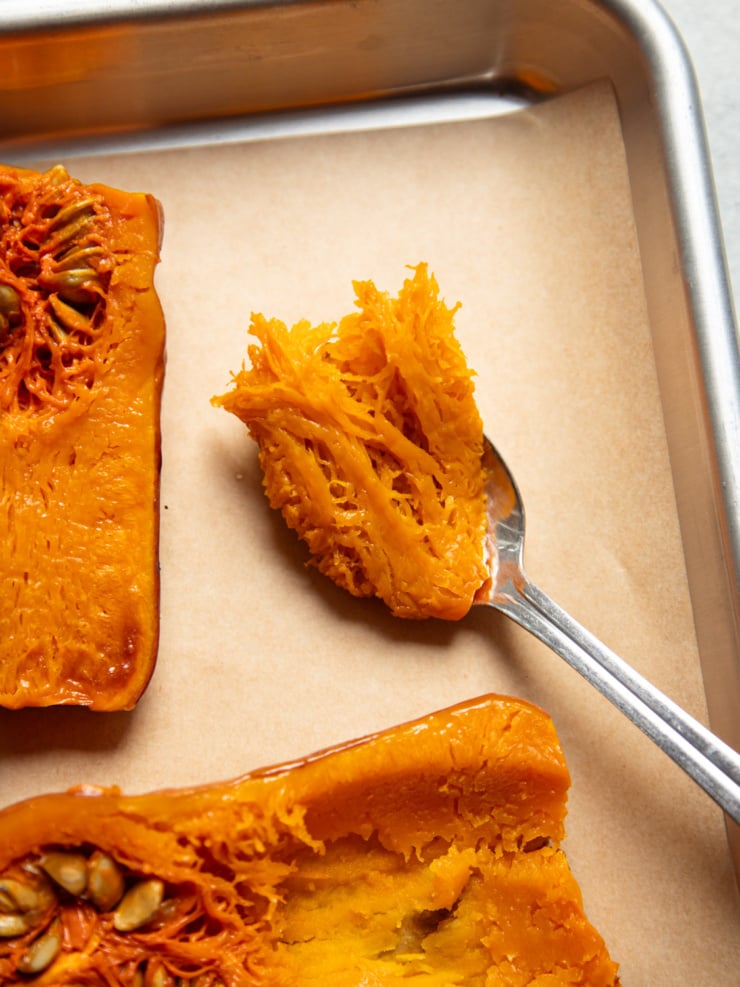 A spoon holds a scoop of stringy pulp from roasted honeynut squash halves on a parchment-lined sheet pan.