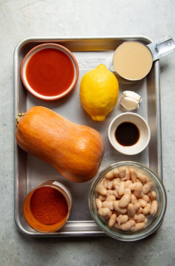 Ingredients for butternut squash dip on a sheet pan: a whole honeynut squash, buffalo hot sauce, lemon, tahini, garlic, vegan Worcestershire, paprika, and white beans.